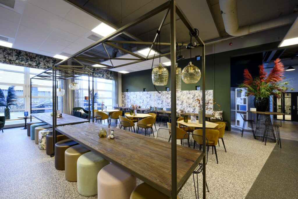 Modern ingerichte kantine aan de H.J.E. Wenckebachweg met lange houten tafels, kleurrijke stoelen en hanglampen.